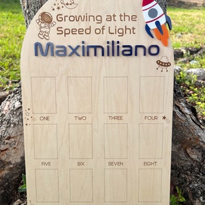 Pu&ograve; includere: Un grafico di crescita in legno a tema spaziale. Presenta un astronauta, un razzo, un UFO e una luna crescente. Il grafico ha 12 spazi per registrare l'altezza di un bambino. Il testo "Growing at the Speed of Light" e "Maximiliano" sono incisi sul grafico.