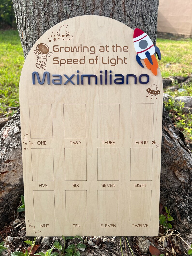 Pu&ograve; includere: Un grafico di crescita in legno a tema spaziale. Presenta un astronauta, un razzo, un UFO e una luna crescente. Il grafico ha 12 spazi per registrare l'altezza di un bambino. Il testo "Growing at the Speed of Light" e "Maximiliano" sono incisi sul grafico.