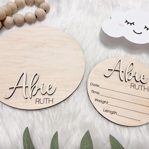 May include: Two wooden round signs with the name "Abie Ruth" in a cursive font. One sign is larger and blank, while the other has lines for date, time, weight, and length. A white cloud decoration with a smiling face is also present.