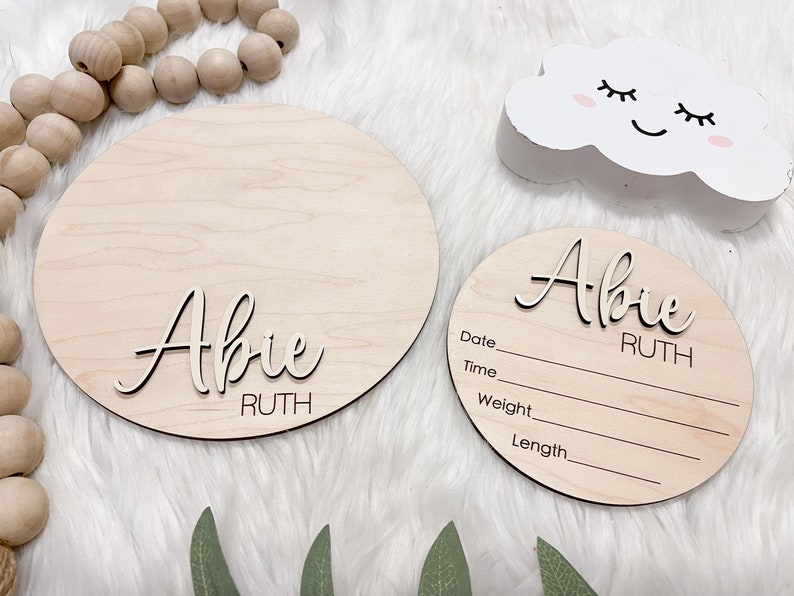 May include: Two wooden round signs with the name "Abie Ruth" in a cursive font. One sign is larger and blank, while the other has lines for date, time, weight, and length. A white cloud decoration with a smiling face is also present.