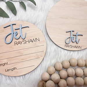 May include: Two round wooden baby announcement signs. One sign has the name "Jet" in blue and "RAYSHAWN" in black. The other sign has the name "Jet" in blue and "RAYSHAWN" in black. Both signs have lines for date, time, weight, and length.