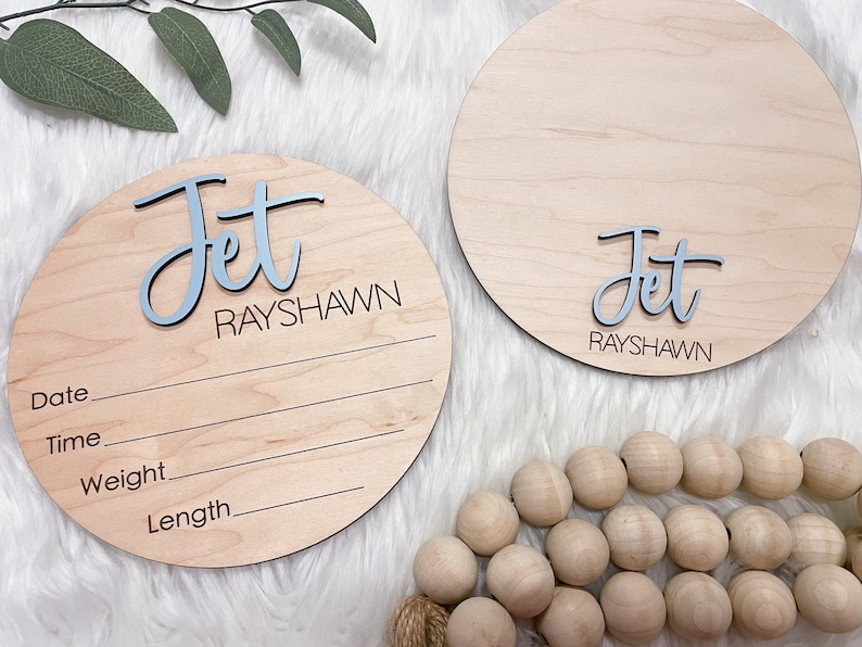 May include: Two round wooden baby announcement signs. One sign has the name "Jet" in blue and "RAYSHAWN" in black. The other sign has the name "Jet" in blue and "RAYSHAWN" in black. Both signs have lines for date, time, weight, and length.