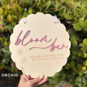 Pu&ograve; includere: Un cartello di legno con un bordo a festoni e il testo "Bloom Bar" in lettere rosa. Il cartello include anche il testo "Please make a bouquet to take on your way".