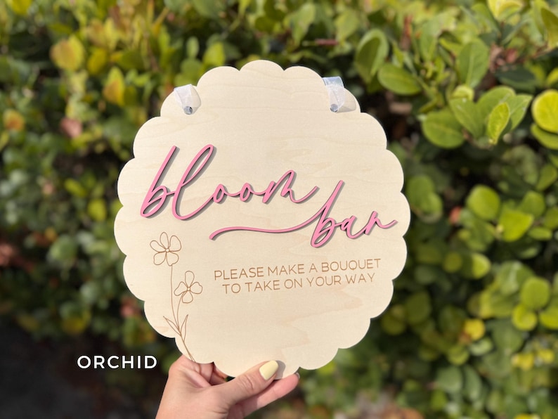 Pu&ograve; includere: Un cartello di legno con un bordo a festoni e il testo "Bloom Bar" in lettere rosa. Il cartello include anche il testo "Please make a bouquet to take on your way".
