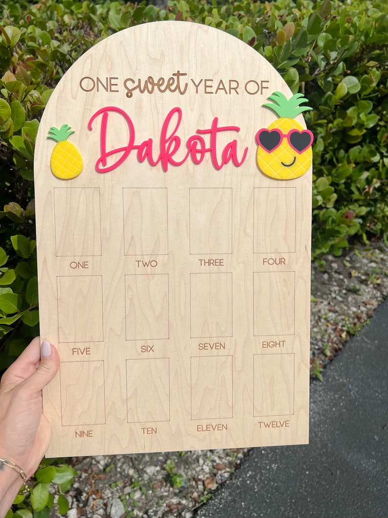 May include: A wooden sign with a pink inscription "ONE sweet YEAR OF Dakota" and a pineapple graphic. The sign has 12 blank squares labeled with numbers one through twelve.