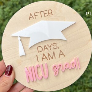 May include: A wooden round sign with the text "After days, I am a NICU grad!" in pink and brown lettering. A white graduation cap is on top of the sign.