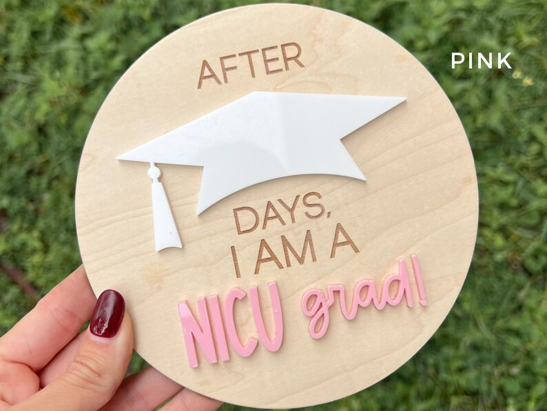 May include: A wooden round sign with the text "After days, I am a NICU grad!" in pink and brown lettering. A white graduation cap is on top of the sign.