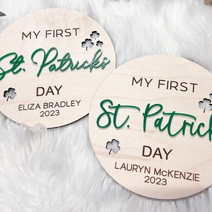 May include: Two round wooden signs with the text "My First St. Patrick's Day" in green and brown lettering. The top sign says "Eliza Bradley 2023" and the bottom sign says "Lauryn McKenzie 2023".