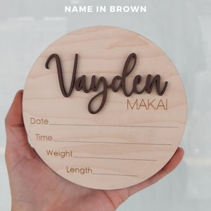 May include: A circular light-colored wooden plaque featuring the name Vayden in raised dark brown script lettering and the name MAKAI in brown text. Below, the words Date, Time, Weight, and Length are printed with horizontal lines for writing.