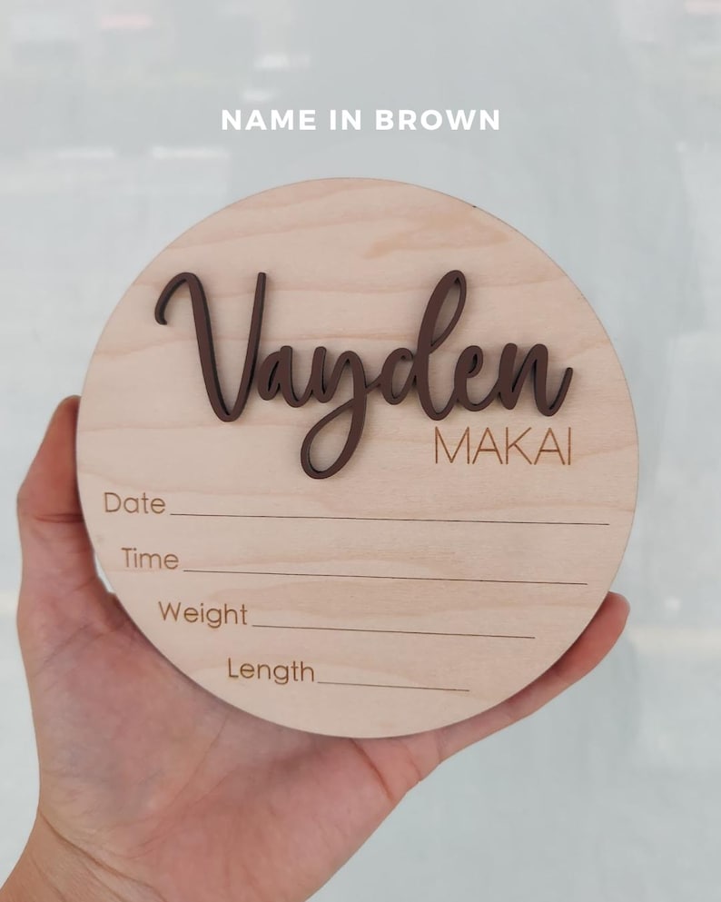 May include: A circular light-colored wooden plaque featuring the name Vayden in raised dark brown script lettering and the name MAKAI in brown text. Below, the words Date, Time, Weight, and Length are printed with horizontal lines for writing.