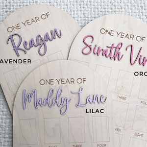 Three birthday wooden boards with arched tops. Each board has a name in a script font and the words "ONE YEAR OF". The boards are labeled "Lavender", "Orchid", and "Lilac". Each board has spaces for photos.