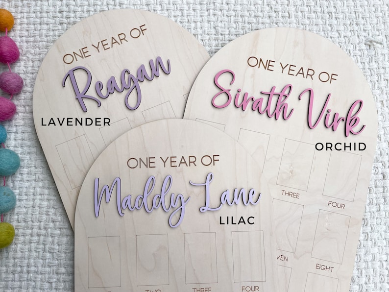 Three birthday wooden boards with arched tops. Each board has a name in a script font and the words "ONE YEAR OF". The boards are labeled "Lavender", "Orchid", and "Lilac". Each board has spaces for photos.