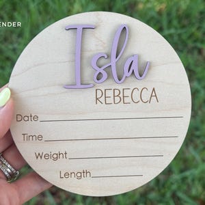A round wooden baby announcement sign with the name "Isla" in lavender script.