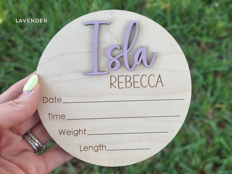 A round wooden baby announcement sign with the name "Isla" in lavender script.
