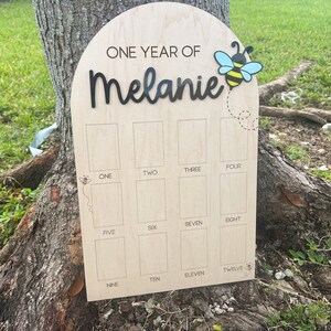 Op de afbeelding: Een boogvormig houten mijlpaalbord met de tekst ONE YEAR OF Melanie in zwarte letters. Het bord bevat twaalf rechthoekige fotovakken gelabeld met de cijfers &eacute;&eacute;n tot en met twaalf, versierd met kleine illustraties van bijen.