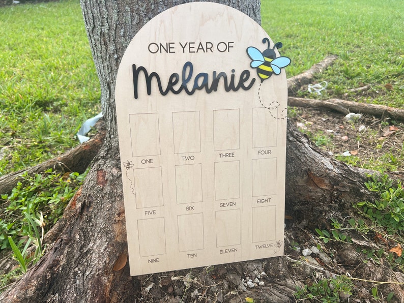Op de afbeelding: Een boogvormig houten mijlpaalbord met de tekst ONE YEAR OF Melanie in zwarte letters. Het bord bevat twaalf rechthoekige fotovakken gelabeld met de cijfers &eacute;&eacute;n tot en met twaalf, versierd met kleine illustraties van bijen.