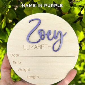 May include: A circular light-colored wooden plaque featuring the name Zoey in raised purple script lettering and the name ELIZABETH in engraved text. Below, the plaque includes engraved lines for Date, Time, Weight, and Length information.