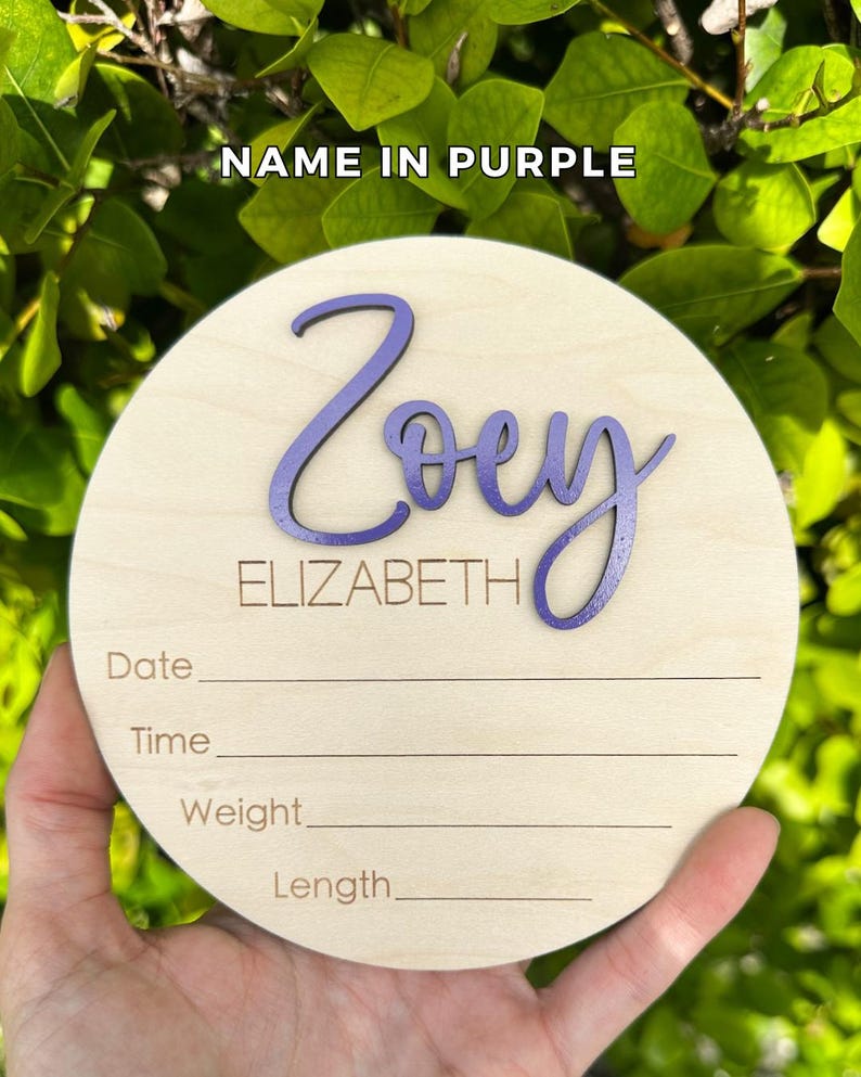 May include: A circular light-colored wooden plaque featuring the name Zoey in raised purple script lettering and the name ELIZABETH in engraved text. Below, the plaque includes engraved lines for Date, Time, Weight, and Length information.