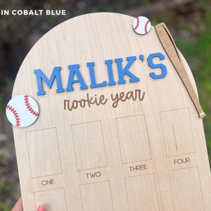 May include: A light-colored wooden plaque with an arched top featuring the text "MALIK'S rookie year" in blue and brown. It includes two baseball cutouts, a wooden baseball bat, and eight rectangular slots labeled "ONE" through "FOUR".
