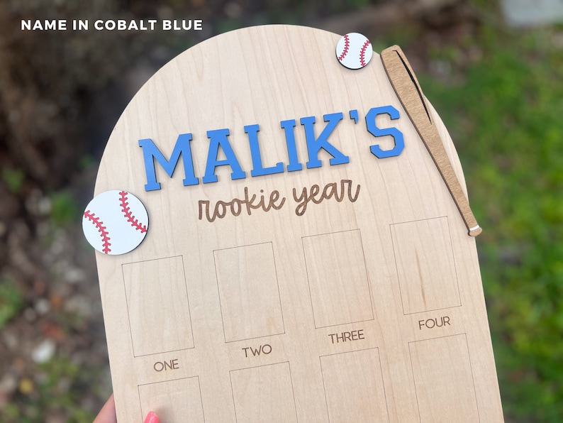 May include: A light-colored wooden plaque with an arched top featuring the text "MALIK'S rookie year" in blue and brown. It includes two baseball cutouts, a wooden baseball bat, and eight rectangular slots labeled "ONE" through "FOUR".