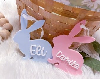 May include: Two bunny-shaped ornaments, one light blue and one pink, with white text. The blue bunny has the name "Eli" and the pink bunny has the name "Camila". Both have white ribbons for hanging. A woven basket is in the background.