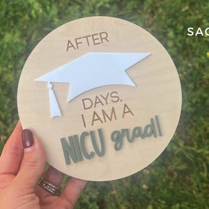 May include: A wooden round sign with the text "After days, I am a NICU grad!" in white and green lettering. A white paper graduation cap is attached to the sign.