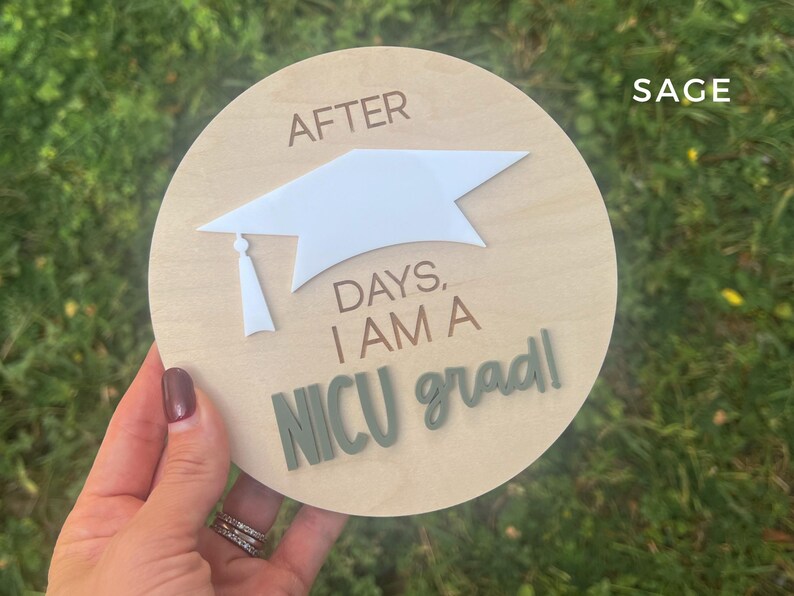 May include: A wooden round sign with the text "After days, I am a NICU grad!" in white and green lettering. A white paper graduation cap is attached to the sign.