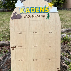 Peut inclure: Un panneau de jalons en bois clair portant le texte KADEN'S first round up. Le panneau comprend douze cadres photo rectangulaires &eacute;tiquet&eacute;s de un &agrave; douze, d&eacute;cor&eacute;s avec des d&eacute;coupes d'un chapeau de cowboy marron, de nuages, d'une &eacute;toile jaune et de figurines vertes.