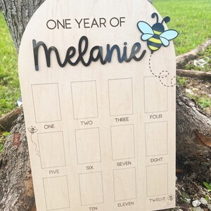 Op de afbeelding: Een boogvormig houten mijlpaalbord met de tekst ONE YEAR OF Melanie in zwarte letters. Een bij-icoon met blauwe en gele vleugels is rechtsboven bevestigd. Twaalf rechthoekige fotovakken zijn gelabeld met de cijfers &eacute;&eacute;n tot en met twaalf.