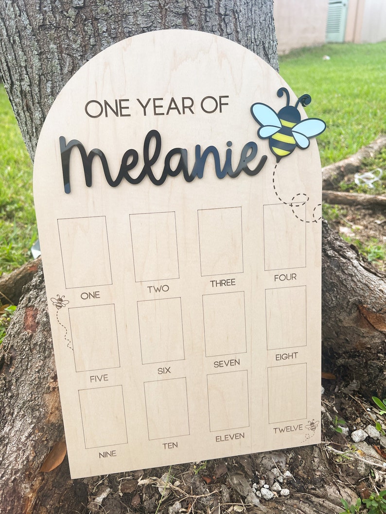 Op de afbeelding: Een boogvormig houten mijlpaalbord met de tekst ONE YEAR OF Melanie in zwarte letters. Een bij-icoon met blauwe en gele vleugels is rechtsboven bevestigd. Twaalf rechthoekige fotovakken zijn gelabeld met de cijfers &eacute;&eacute;n tot en met twaalf.