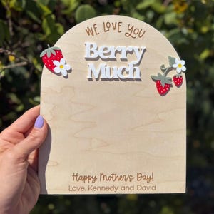 May include: A wooden sign with the words "We Love You Berry Much" and "Happy Mother's Day!" in a script font. The sign is decorated with red strawberries and white flowers. The sign is held by a person.