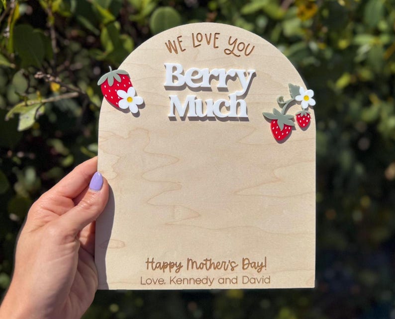 May include: A wooden sign with the words "We Love You Berry Much" and "Happy Mother's Day!" in a script font. The sign is decorated with red strawberries and white flowers. The sign is held by a person.