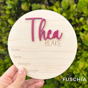 May include: A circular wooden birth announcement sign featuring the name Thea in raised fuchsia script lettering and the name BLAKE in engraved text. Below the names are four engraved lines with the labels Date, Time, Weight, and Length.