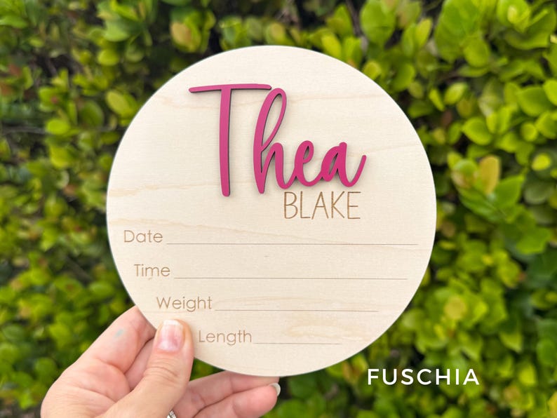 May include: A circular wooden birth announcement sign featuring the name Thea in raised fuchsia script lettering and the name BLAKE in engraved text. Below the names are four engraved lines with the labels Date, Time, Weight, and Length.