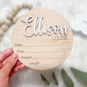 May include: A round wooden birth announcement sign with the name "Ellison" in white script, and "Louise" in a smaller font. The sign has lines for date, time, weight, and length. A wooden bead garland and a cloud decoration are in the background.