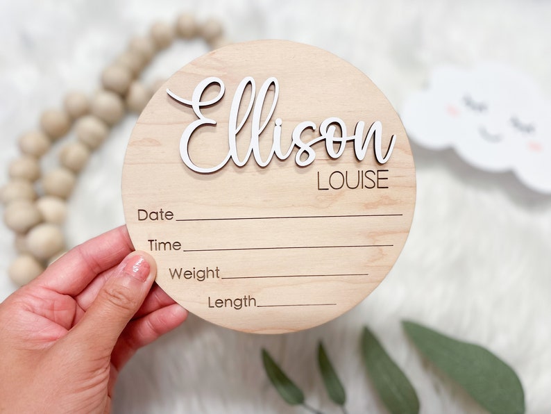 May include: A round wooden birth announcement sign with the name "Ellison" in white script, and "Louise" in a smaller font. The sign has lines for date, time, weight, and length. A wooden bead garland and a cloud decoration are in the background.
