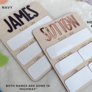 May include: Two rectangular wooden milestone boards with rounded corners. One features the name JAMES in navy blue, the other SUTTON in metallic rose. Both include white dry-erase sections with text: TODAY I AM, I REALLY LOVE, I CAN, I DON'T LIKE, and MORE ABOUT ME.
