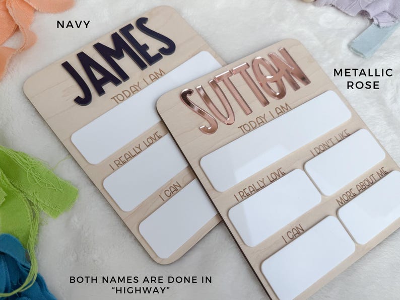 May include: Two rectangular wooden milestone boards with rounded corners. One features the name JAMES in navy blue, the other SUTTON in metallic rose. Both include white dry-erase sections with text: TODAY I AM, I REALLY LOVE, I CAN, I DON'T LIKE, and MORE ABOUT ME.