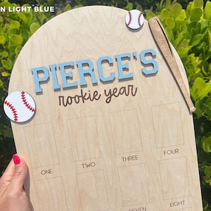 May include: A light-colored wooden plaque shaped like home plate featuring the text PIERCE'S rookie year in light blue and brown. The plaque includes two baseball cutouts, a wooden bat cutout, and twelve rectangular slots labeled one through twelve.