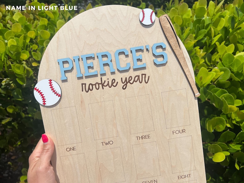 May include: A light-colored wooden plaque shaped like home plate featuring the text PIERCE'S rookie year in light blue and brown. The plaque includes two baseball cutouts, a wooden bat cutout, and twelve rectangular slots labeled one through twelve.
