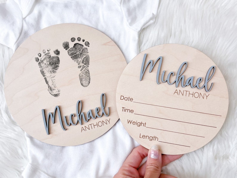 May include: Two round wooden baby announcement plaques. One features baby footprints and the name "Michael Anthony" in gray script. The other has the same name and lines for date, time, weight, and length.