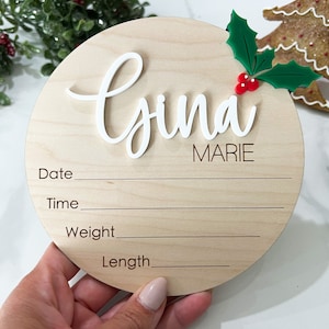 May include: A round wooden plaque with the name "Gyna Marie" written in white cursive lettering. The plaque has spaces to fill in the date, time, weight, and length. A small green holly sprig with red berries is attached to the top of the plaque.