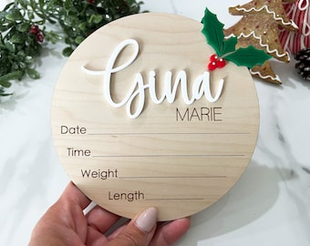 May include: A round wooden plaque with the name "Gyna Marie" written in white cursive lettering. The plaque has spaces to fill in the date, time, weight, and length. A small green holly sprig with red berries is attached to the top of the plaque.