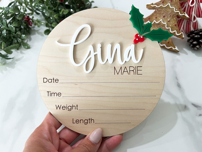 May include: A round wooden plaque with the name "Gyna Marie" written in white cursive lettering. The plaque has spaces to fill in the date, time, weight, and length. A small green holly sprig with red berries is attached to the top of the plaque.