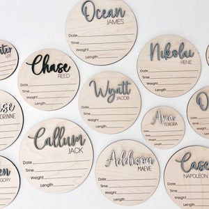 K&ouml;nnte beinhalten: Eine Sammlung runder Baby-Ank&uuml;ndigungsschilder aus Holz. Jedes Schild zeigt einen Namen in einer geschichteten, grau-wei&szlig;en Schrift. Die Schilder enthalten Felder zur Aufzeichnung von Datum, Uhrzeit, Gewicht und L&auml;nge des Babys.