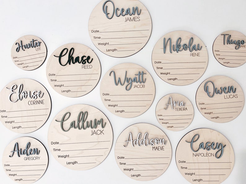 K&ouml;nnte beinhalten: Eine Sammlung runder Baby-Ank&uuml;ndigungsschilder aus Holz. Jedes Schild zeigt einen Namen in einer geschichteten, grau-wei&szlig;en Schrift. Die Schilder enthalten Felder zur Aufzeichnung von Datum, Uhrzeit, Gewicht und L&auml;nge des Babys.