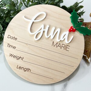 May include: A round wooden plaque with a white cursive name "Gina Marie" and a green holly sprig with red berries. The plaque has lines for recording the date, time, weight, and length.