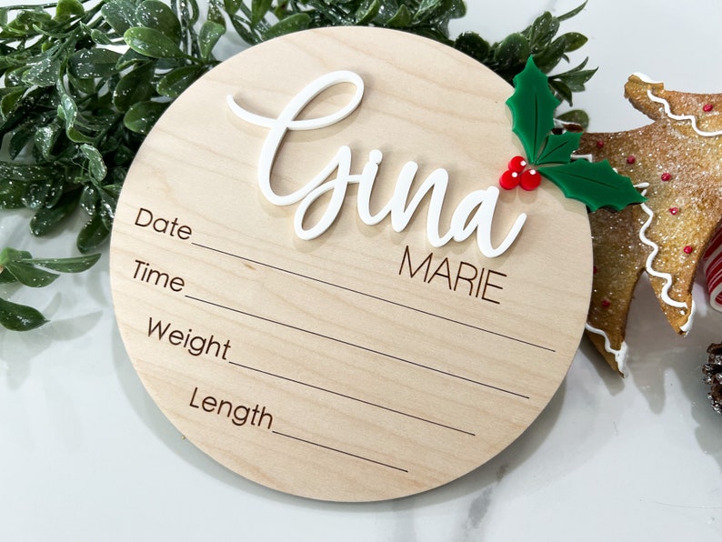 May include: A round wooden plaque with a white cursive name "Gina Marie" and a green holly sprig with red berries. The plaque has lines for recording the date, time, weight, and length.