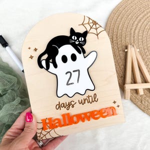 K&ouml;nnte beinhalten: H&ouml;lzernes Halloween-Countdown-Schild mit einer schwarzen Katze auf einem wei&szlig;en Geist, der die Zahl 27 anzeigt. Auf dem Schild steht "Tage bis Halloween" in Orange, mit einem Spinnennetz-Design. Ein schwarzer Marker und eine kleine Holzstaffelei sind ebenfalls sichtbar.