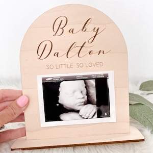 May include: A wooden arch-shaped sign with the text "Baby Dalton So Little So Loved" engraved on it. A black and white ultrasound image is displayed in the centre of the sign.